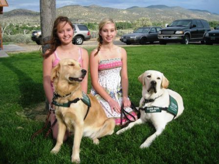 Two girls are sitting next to two dogs one of which has a harness on