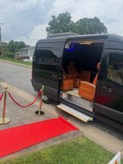 Black van with open side door beside a red carpet and roped walkway on a wet street.