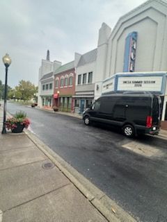 Rainy street with storefronts, a black van parked curbside, and a sidewalk with planters and a lamppost.