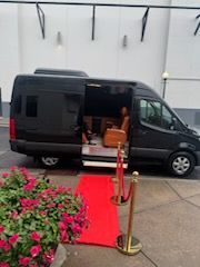 Black van with open side door beside a red carpet entrance and flower arrangements in a driveway.
