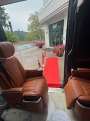 Interior of a luxury van with brown leather seats facing a red carpeted entrance at a building door
