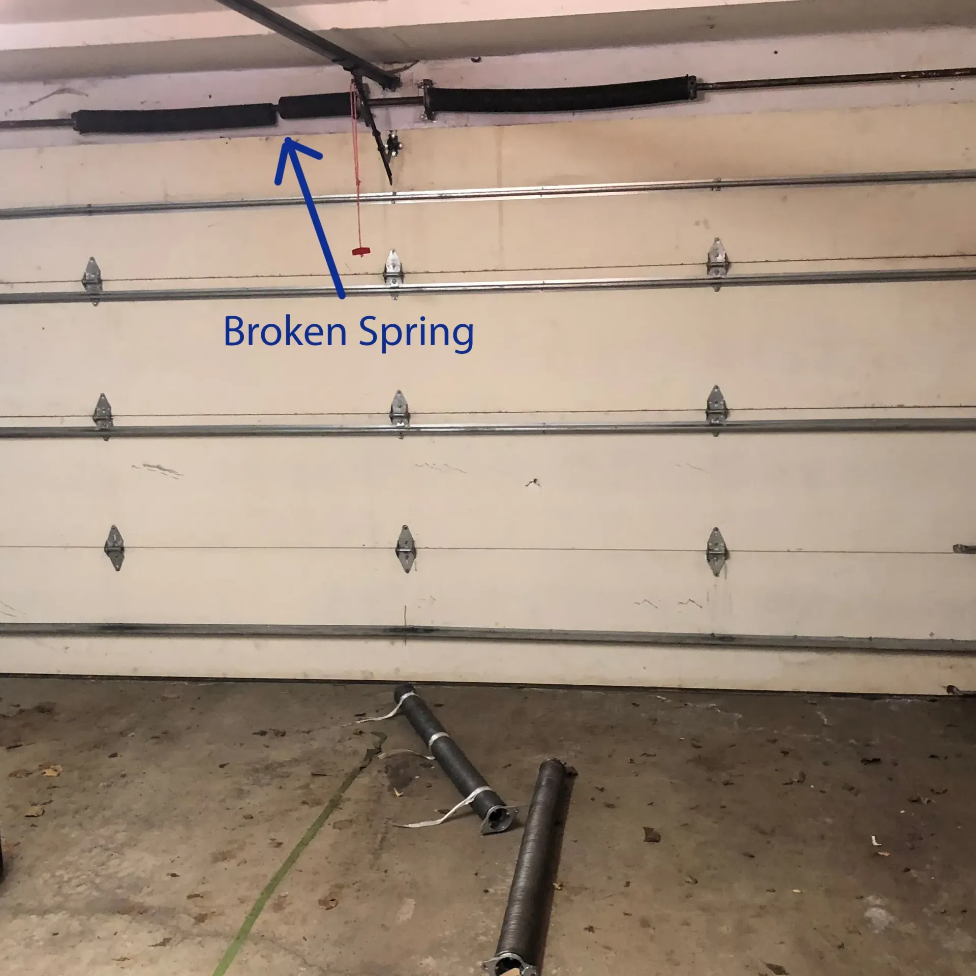 Garage door with a broken spring, shown with an arrow pointing to the damaged area and parts on the floor.