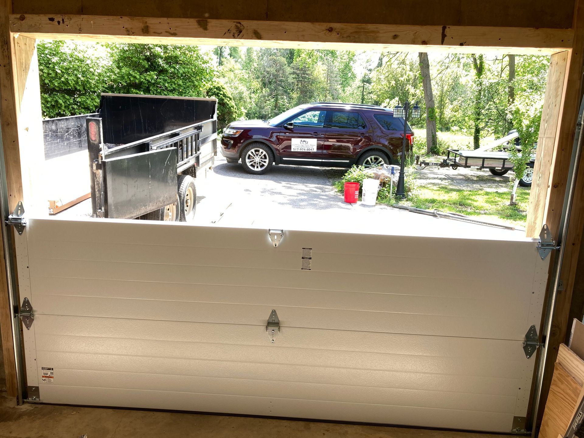 Garage door opens to a driveway with a maroon SUV and a black dumpster in bright daylight
