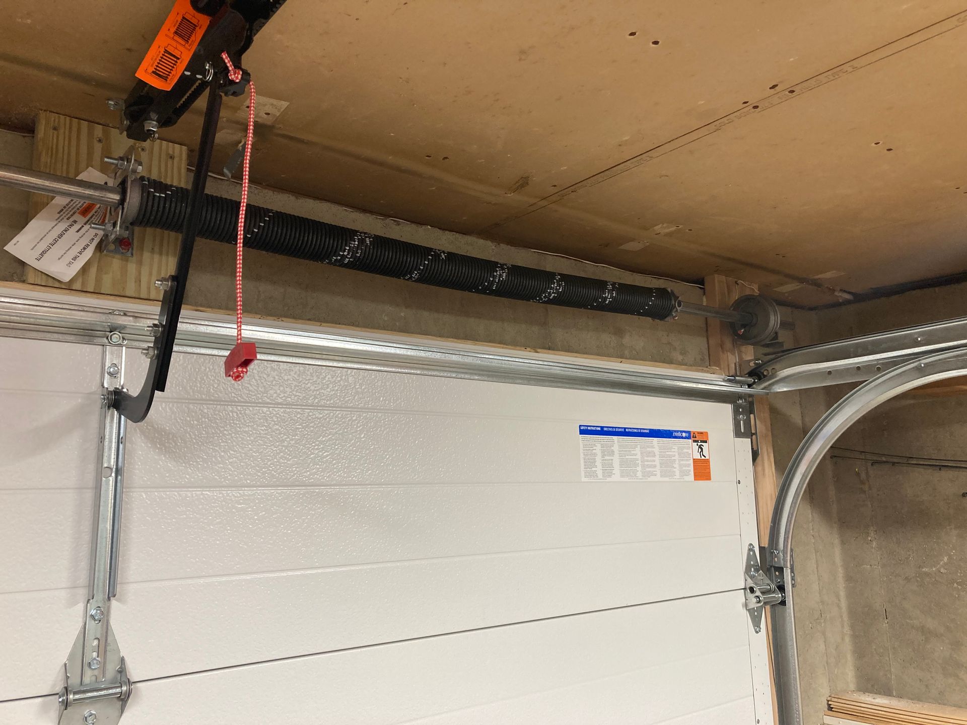 Garage ceiling with a ceiling-mounted orange hoist, cable, and a white garage door below.