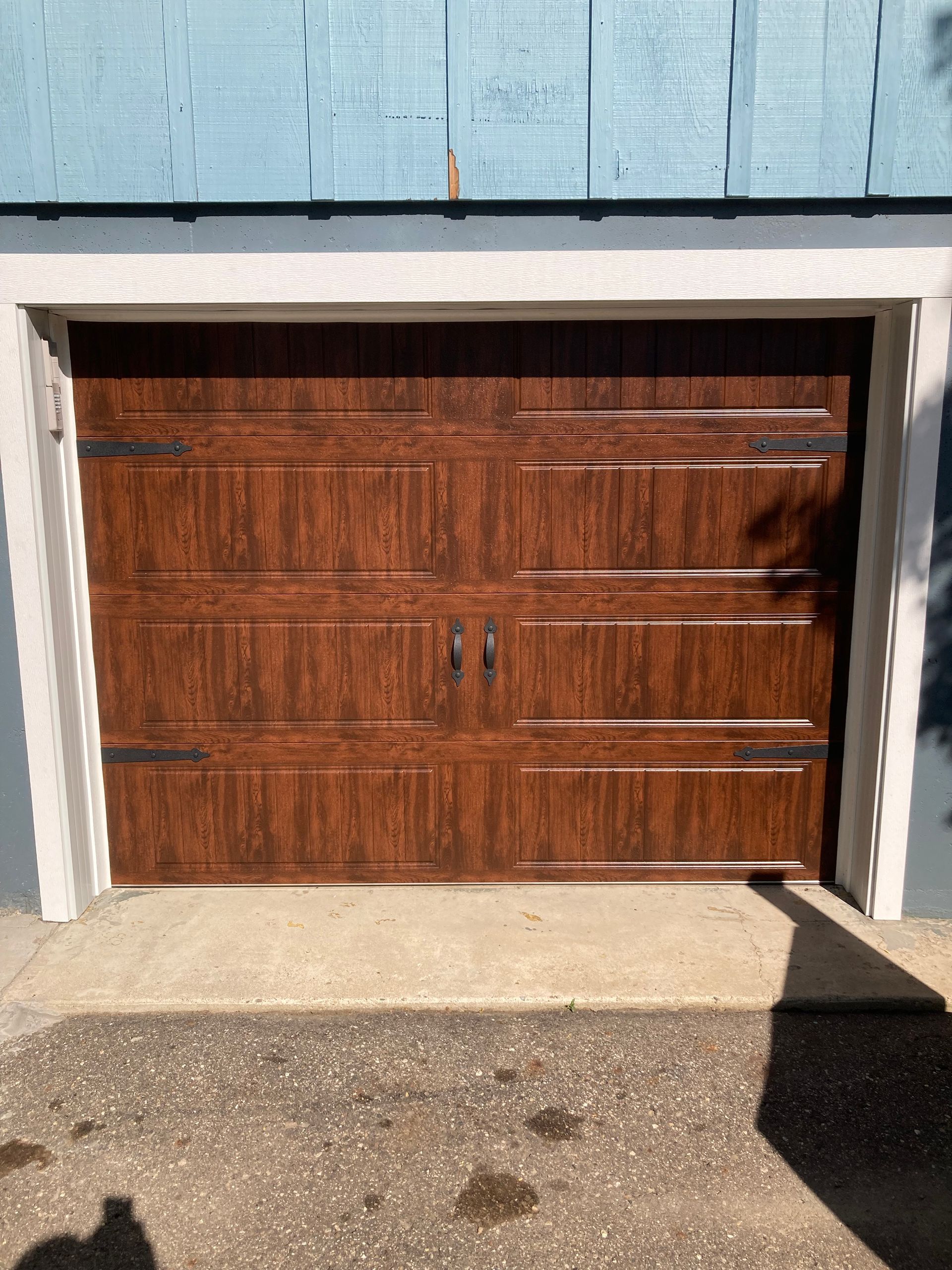Wooden garage door in a white frame beneath blue siding, with concrete driveway in front.