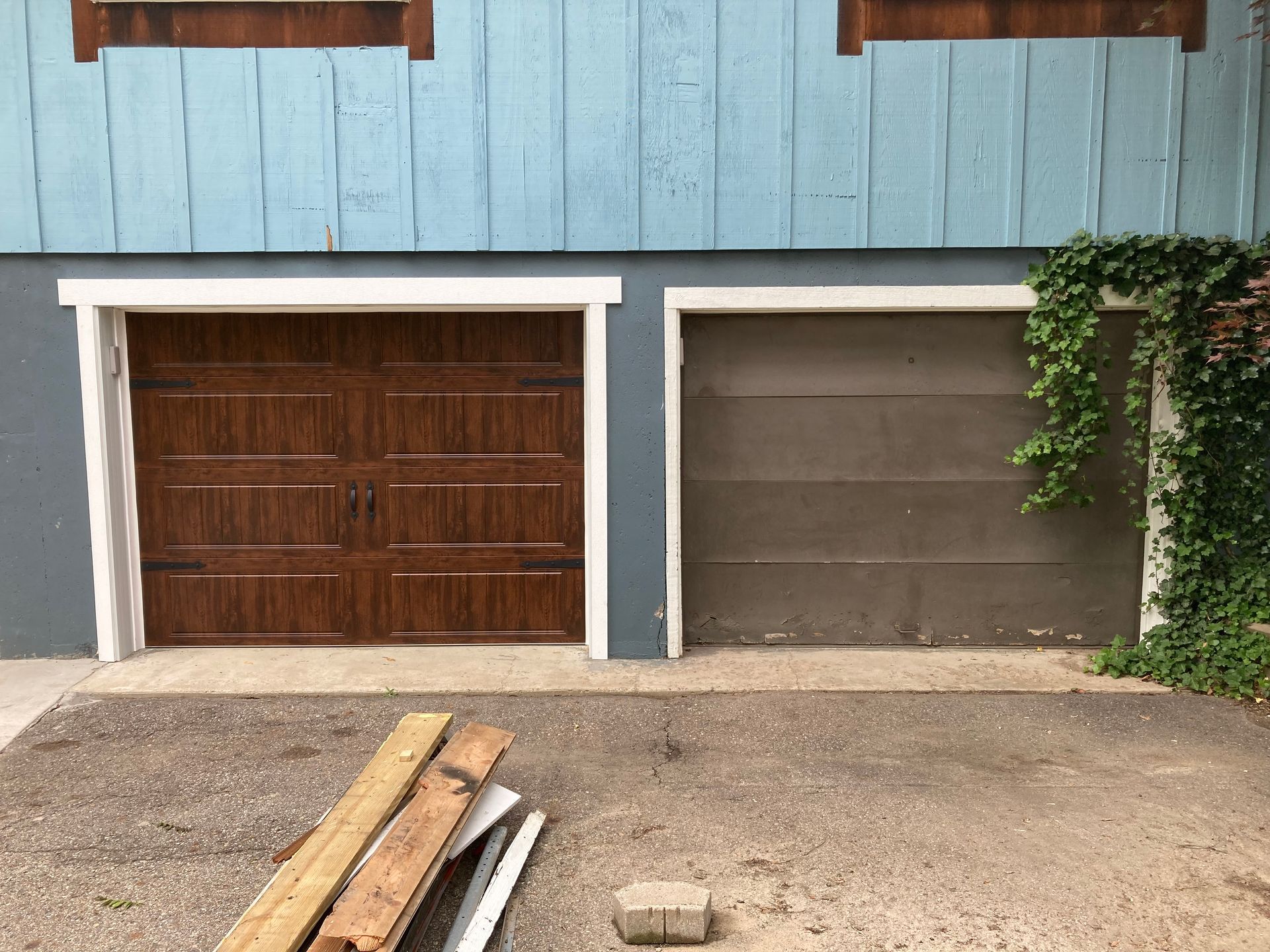 Two garage doors on a house, one brown and one gray, with wood planks on the driveway.