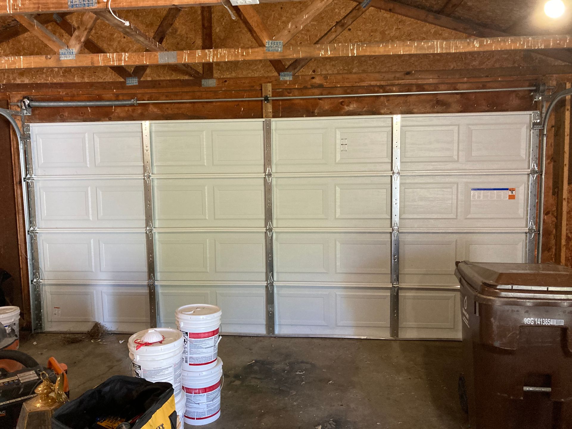 White paneled garage door inside a wood-framed garage with paint buckets and a trash bin nearby