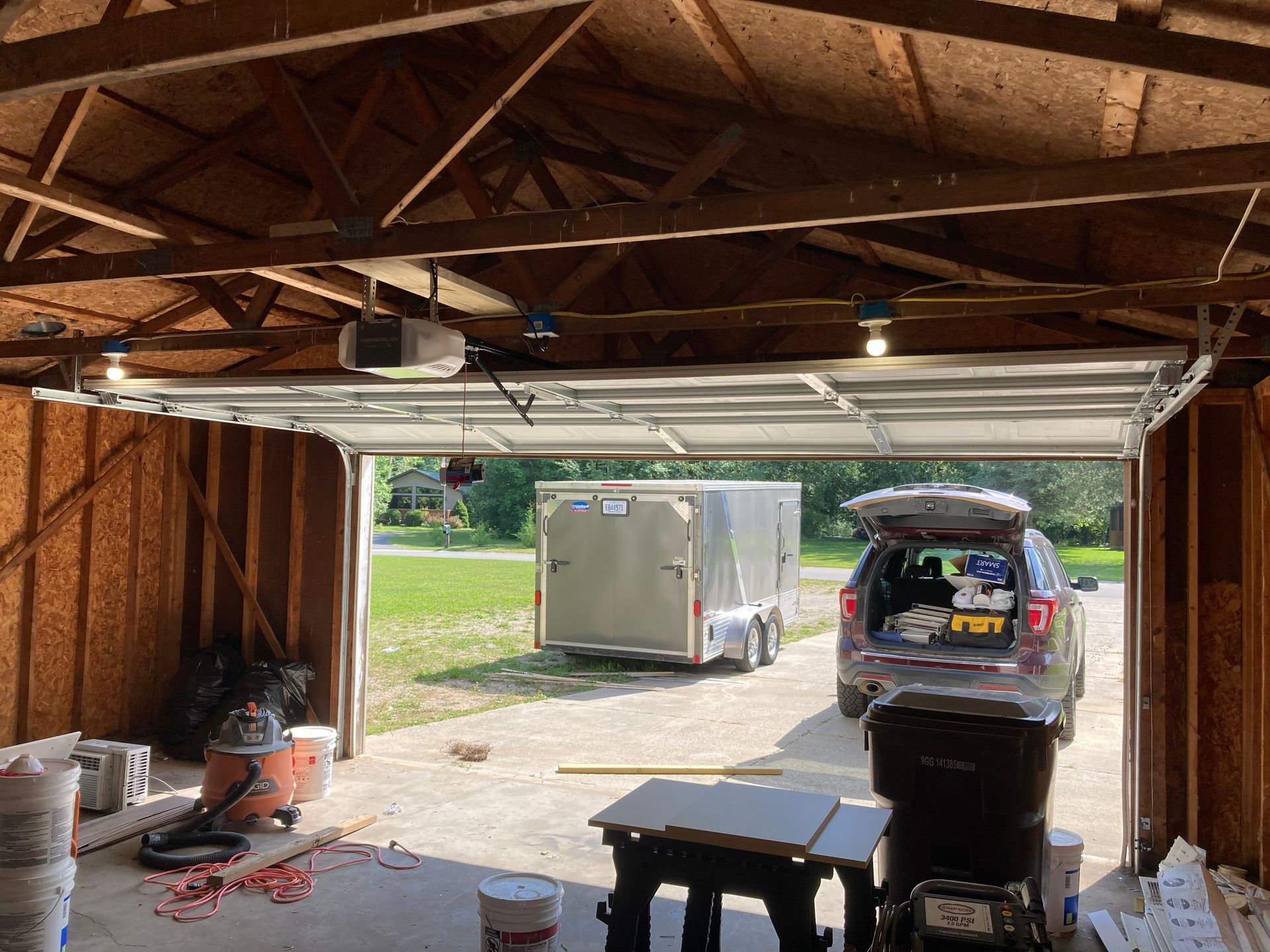Open garage with a trailer outside and a hatchback SUV loaded with items, workbench and tools inside.