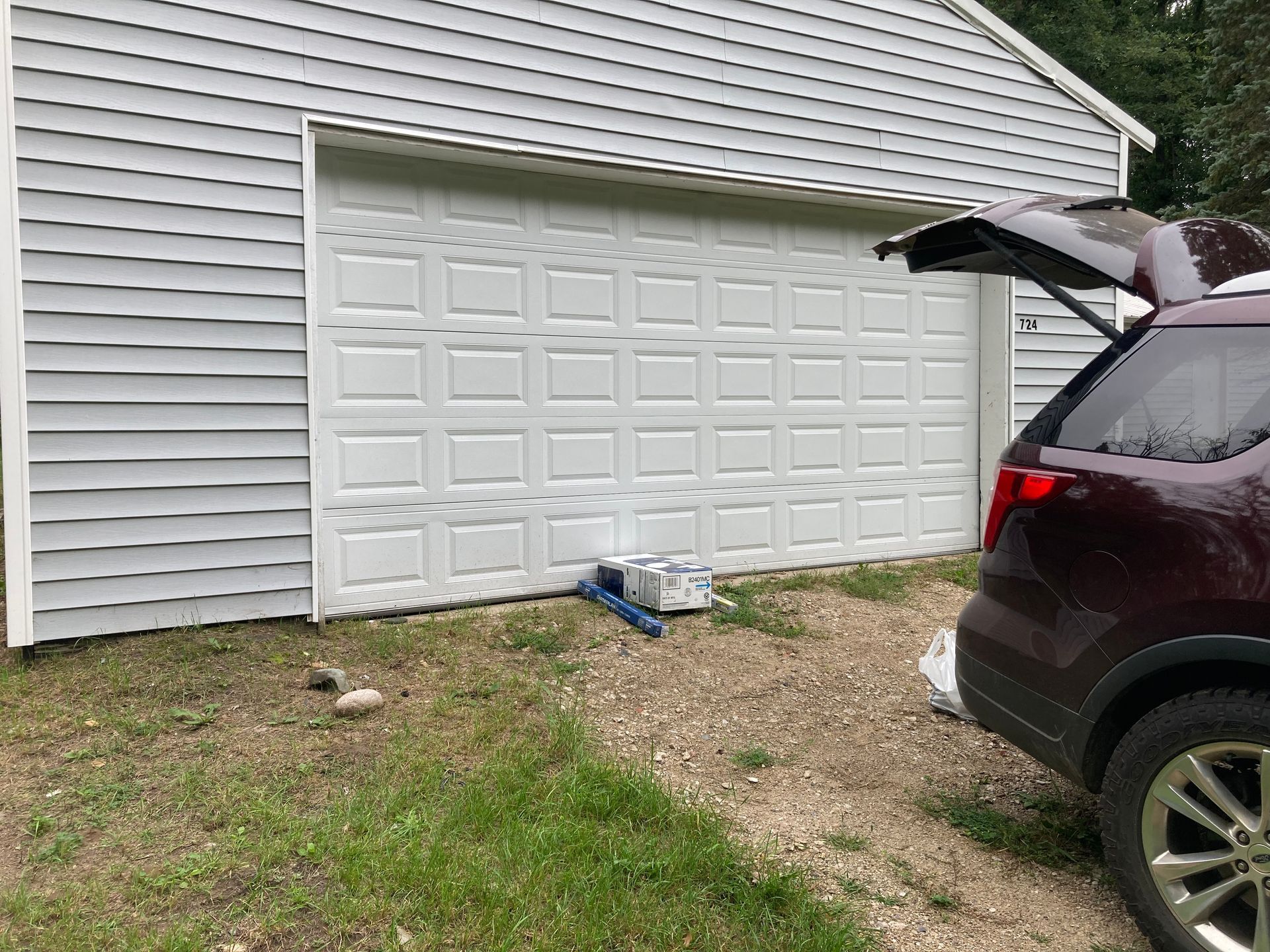 Suburban garage with white overhead door, a maroon SUV parked beside it, and a cordless tool on the ground.