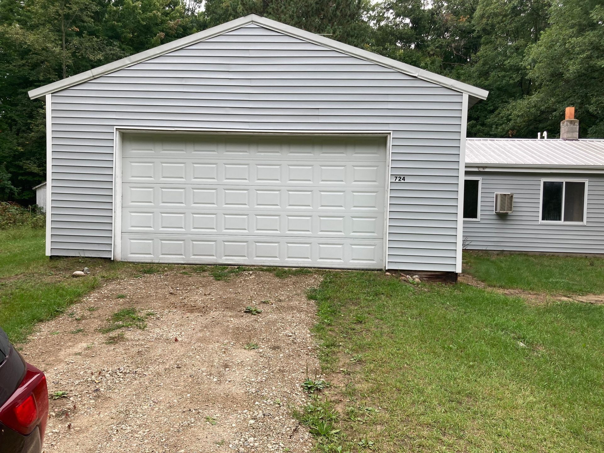 White garage with double door and gravel driveway beside a small house, bordered by grass and trees