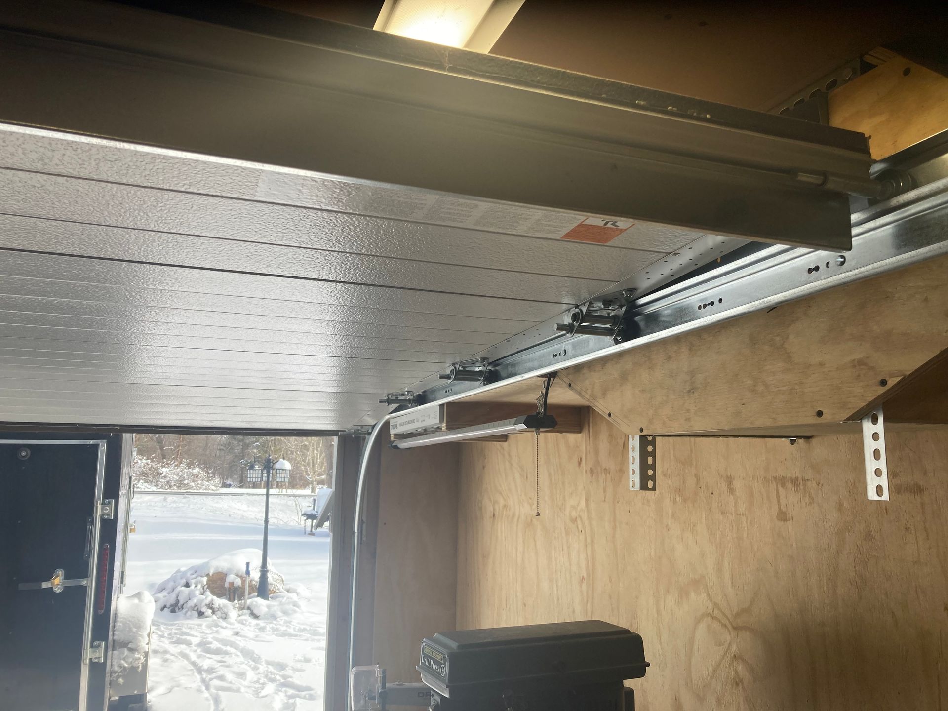 Garage interior with an open door, overhead track, and plywood walls; snowy yard visible outside