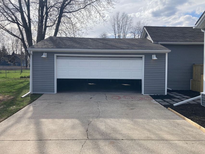 Detached gray garage with white door beside a driveway and house under a cloudy sky