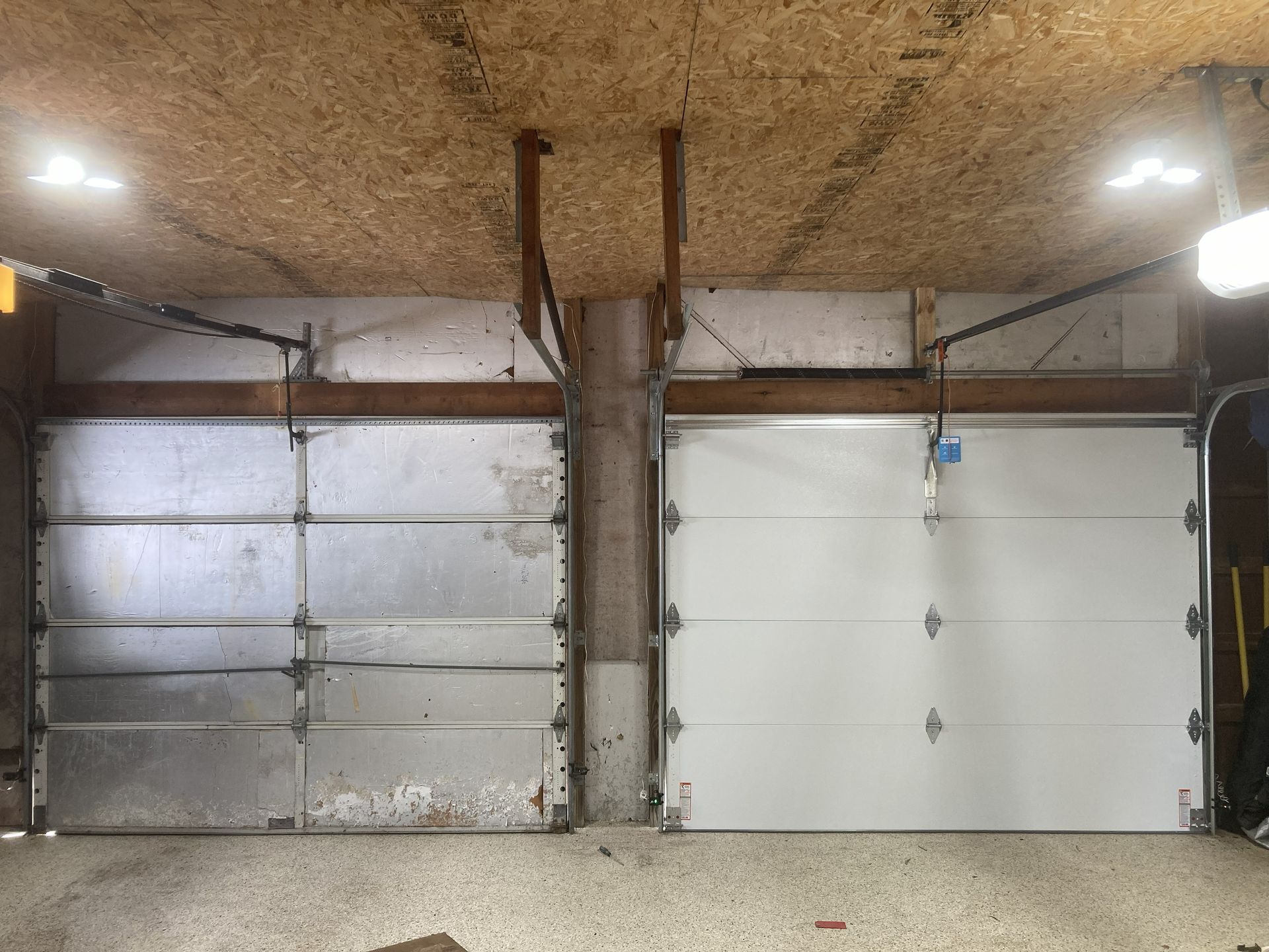 Three garage doors in a concrete garage, with two silver doors and one white door under a low ceiling.
