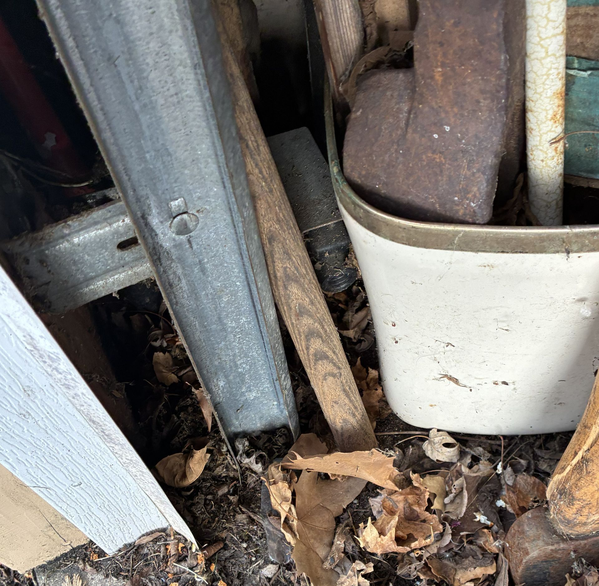 Rusty metal and wooden yard tools stored in a white plastic bin among leaves and debris.