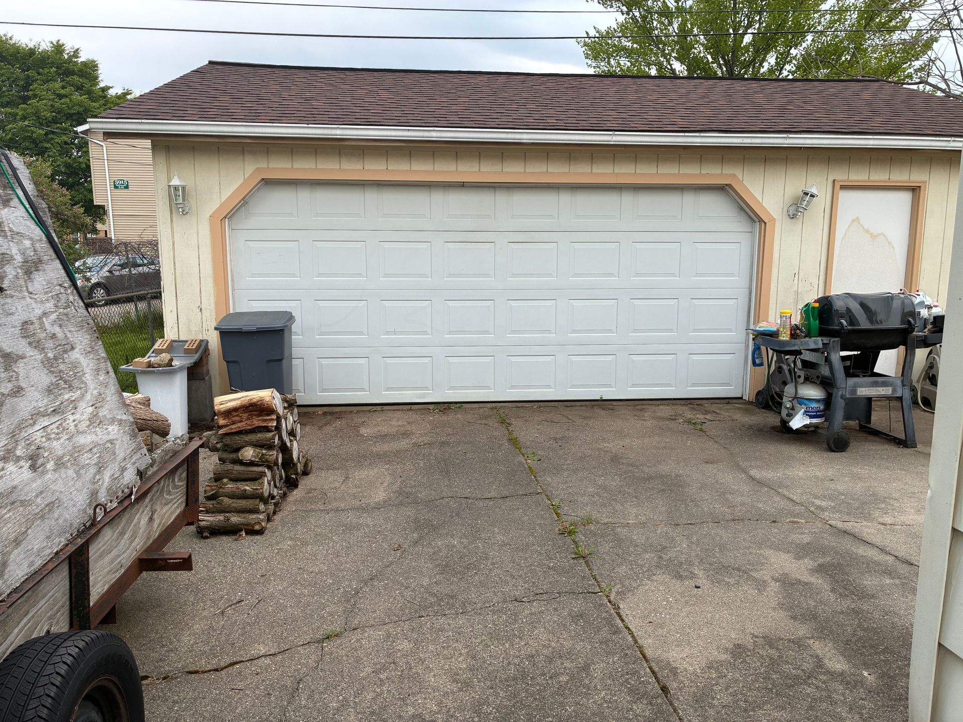 Detached garage with white door, cracked concrete driveway, and stacked firewood beside a worktable