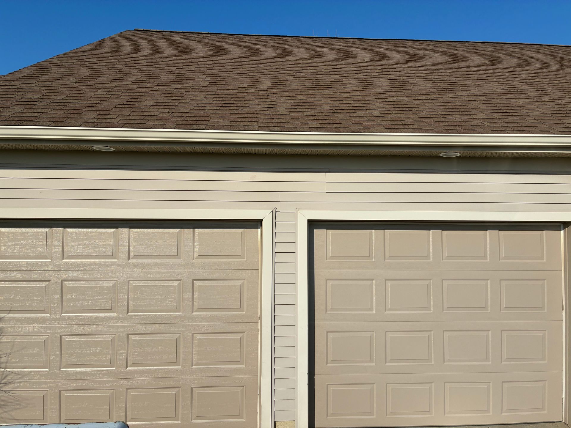 Two beige garage doors beneath a brown shingled roof on a house exterior