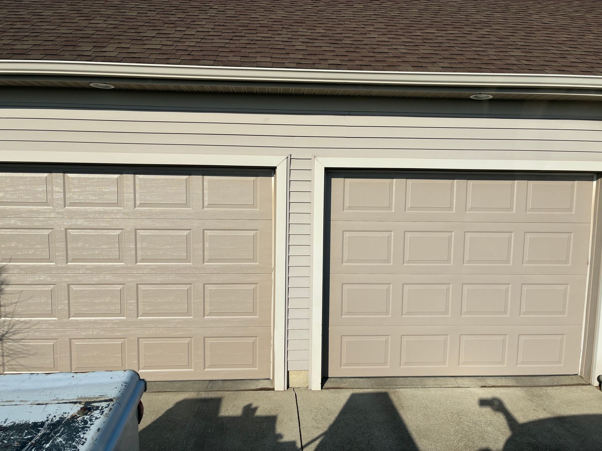 Two beige garage doors on a house exterior, with a partial blue car at left and snow on the driveway.
