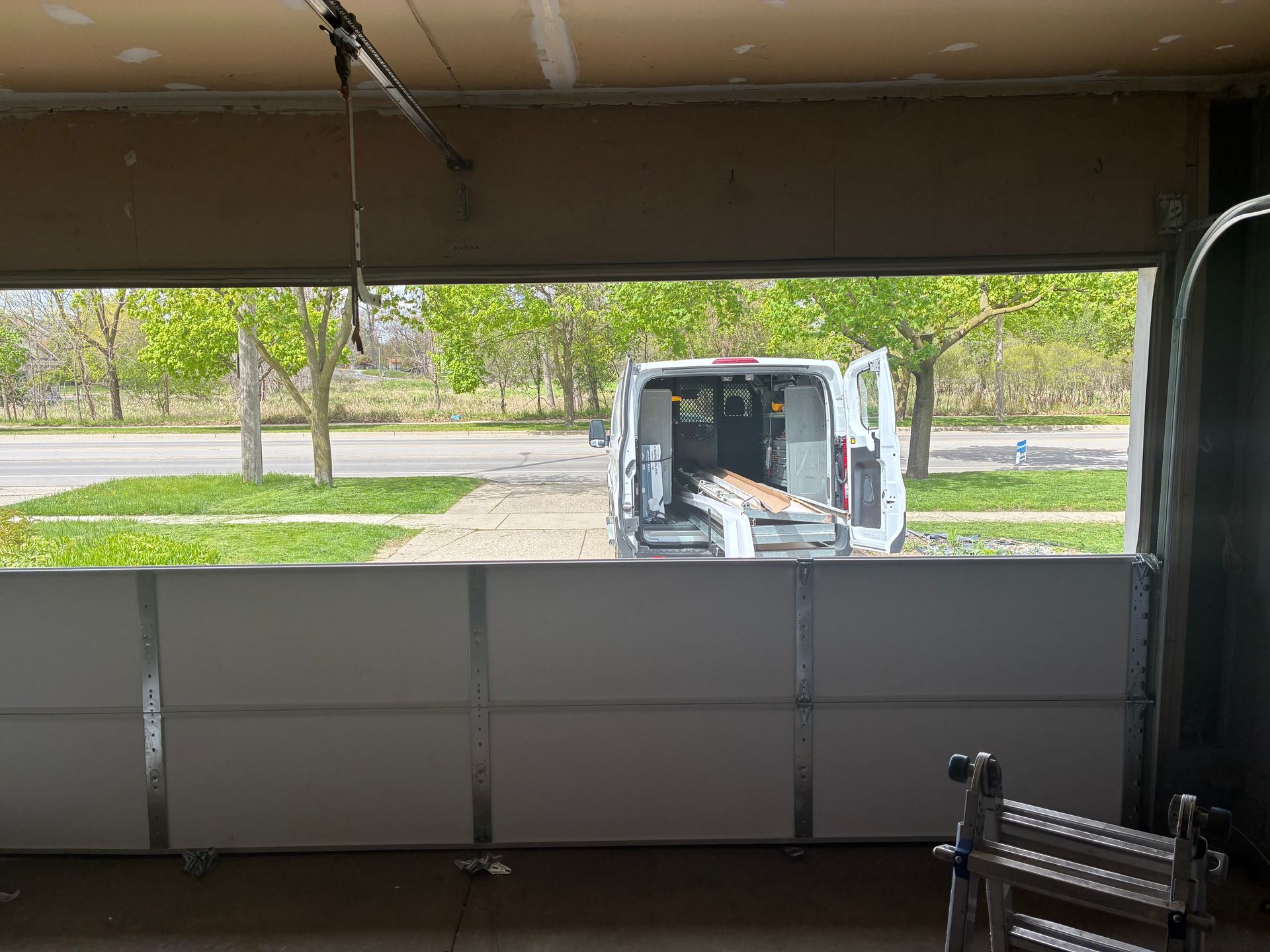 Inside a garage, a white utility truck is parked outside a wide open door, with trees beyond.