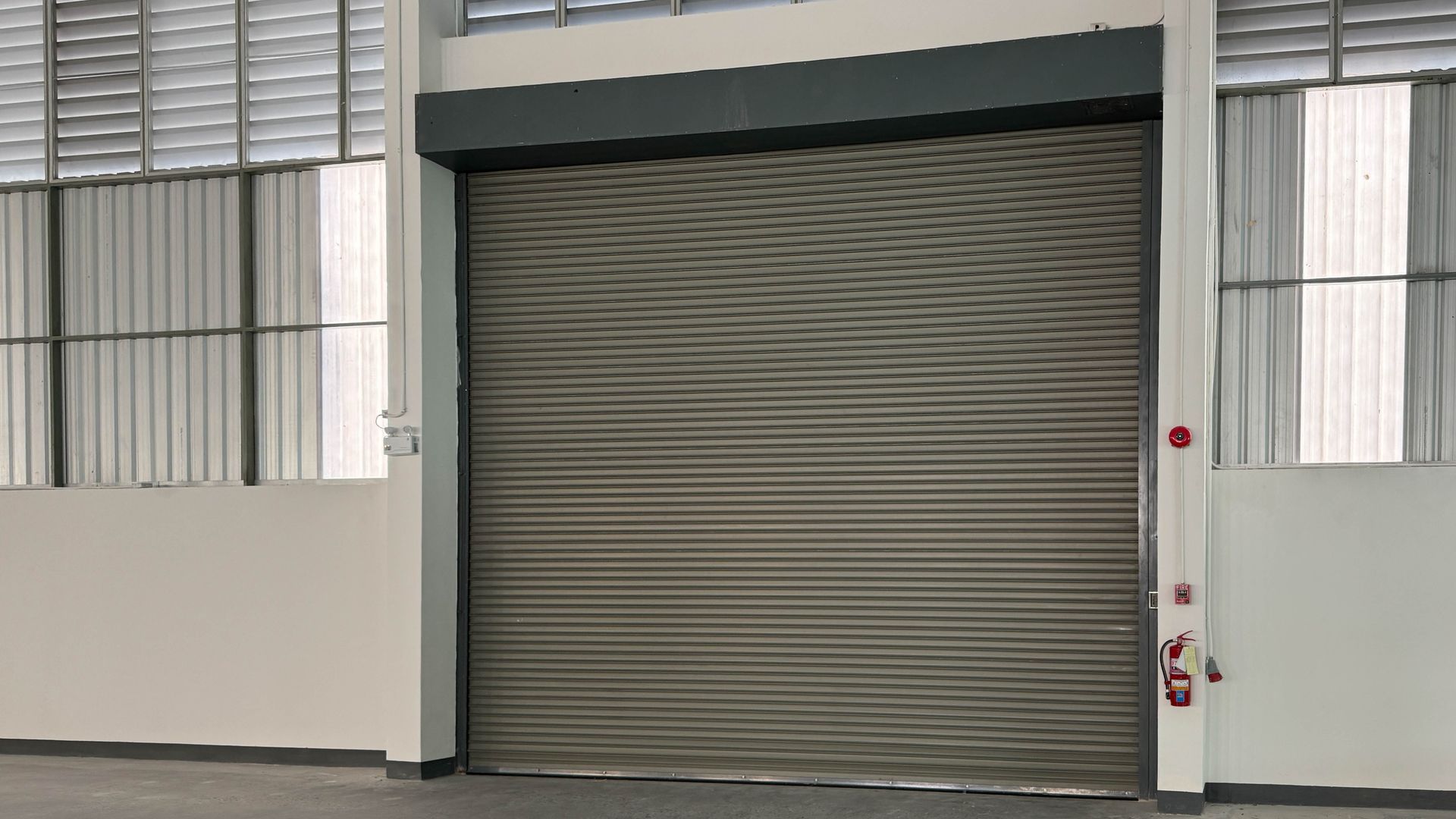 Closed gray metal roll-up door in a building interior, with white walls and narrow windows on both sides