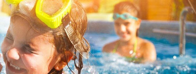 Two children wearing swimming goggles smile while playing in a sunny pool.