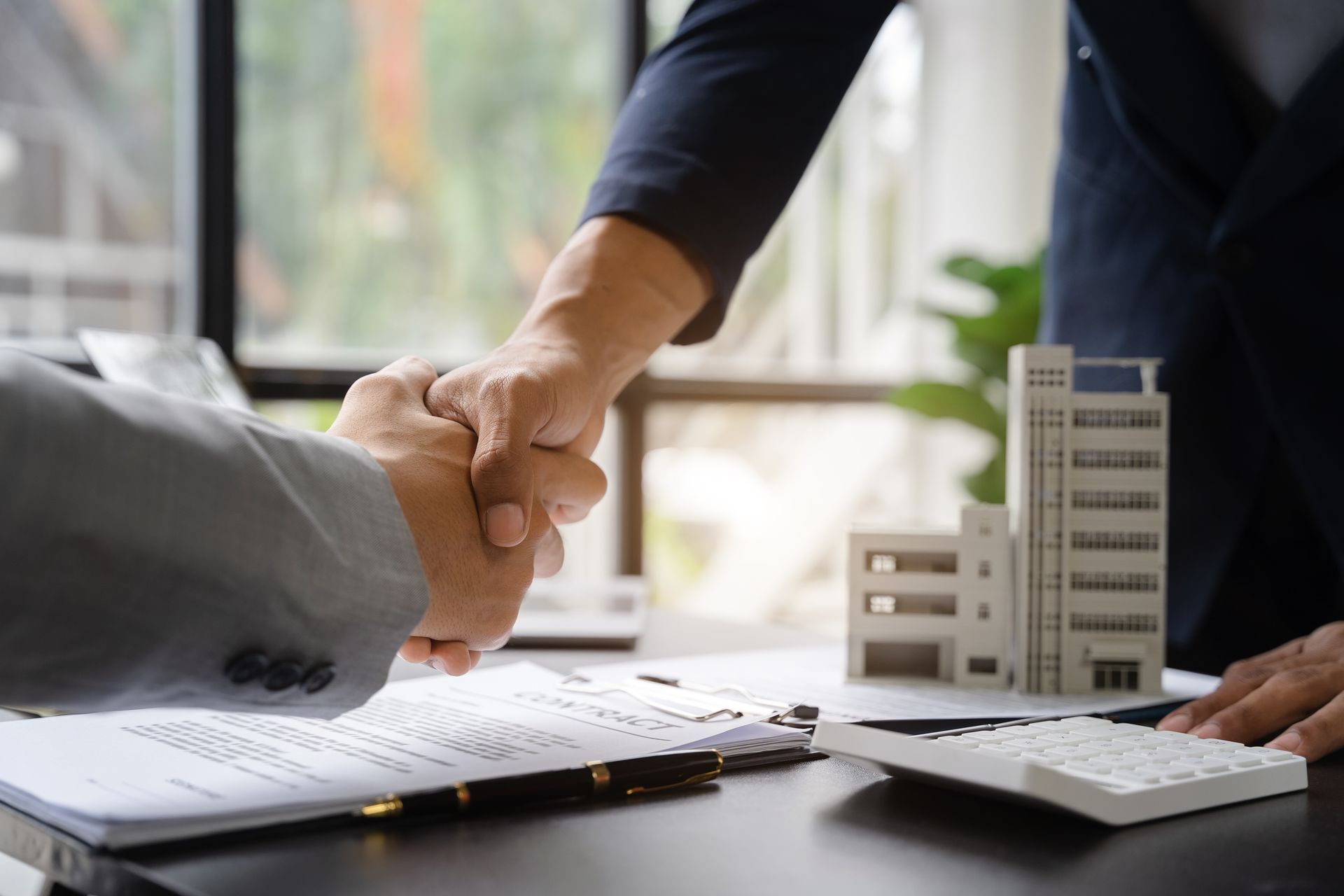 Businesspeople shaking hands over a desk with documents, calculator, and model building in an office