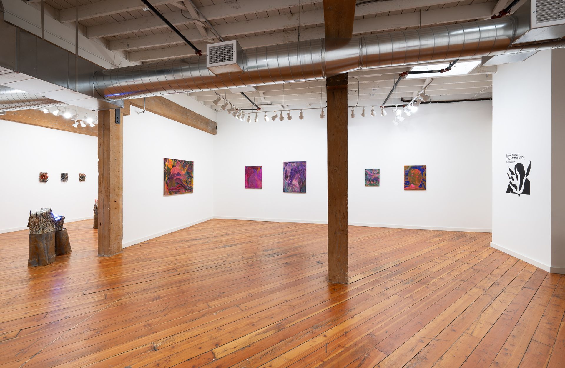 An art gallery space with wooden floors, white walls hung with colorful paintings by Emily Elizabeth Wise, and two pillars.