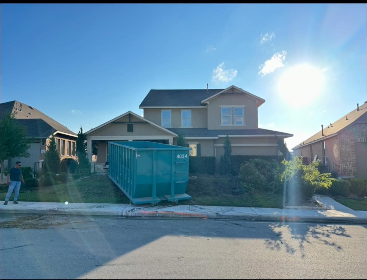 A green dumpster sits in the driveway of a two-story house, likely for renovation. Sunny day.