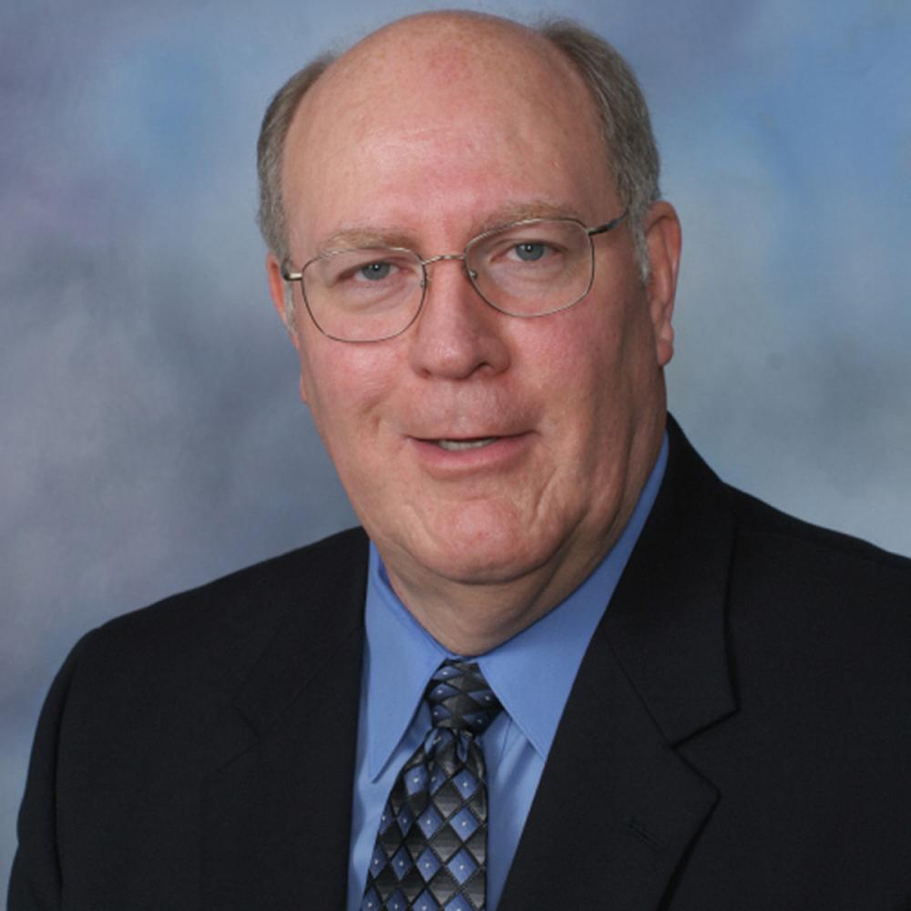 Bald man in suit and glasses, smiling at the camera. Blue and black patterned tie, against a blue background.
