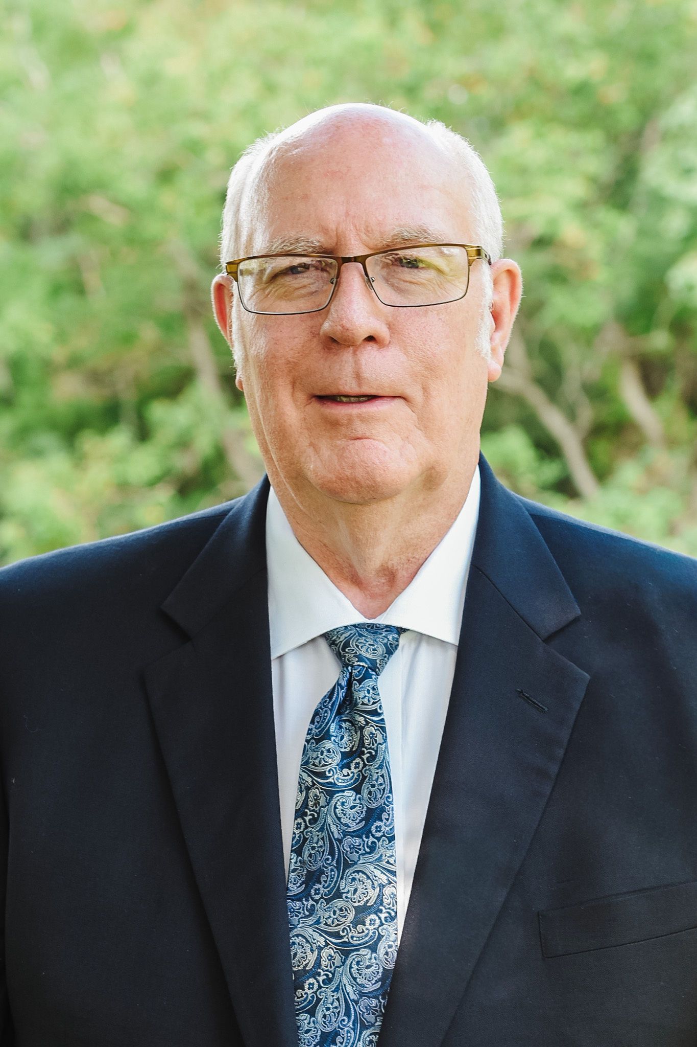 Bald man in suit and glasses, smiling at the camera. Blue and black patterned tie, against a blue background.