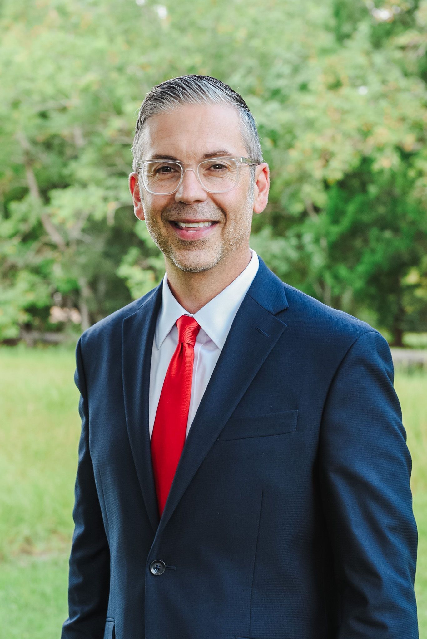 Man in suit and glasses smiling in front of green trees.