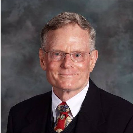 Older man with glasses, wearing a suit and tie, smiling against a blurry background.