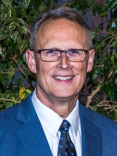 Older man with glasses, wearing a suit and tie, smiling against a blurry background.