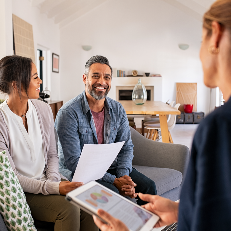 Three people chatting on a couch in a bright living room, holding papers and smiling.