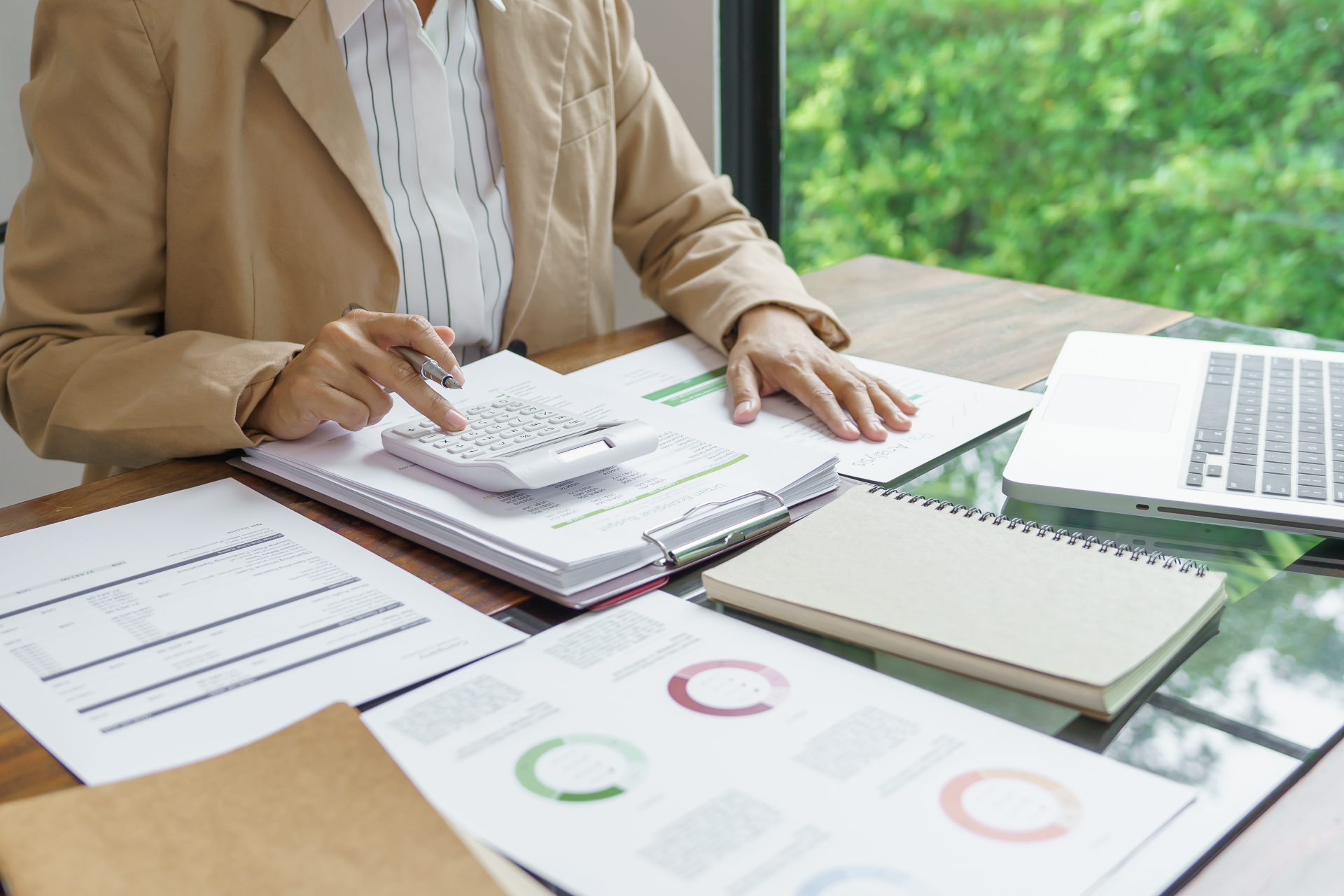Person taking notes at a desk with laptop, notebook, papers, and charts in a bright office.
