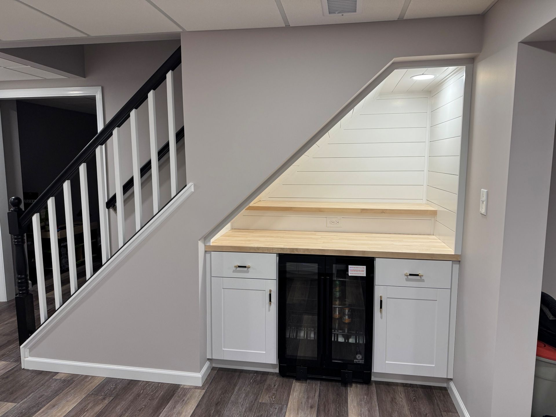 A white bar nook with a wooden countertop and wine fridge built into the empty space under a staircase with a black railing.