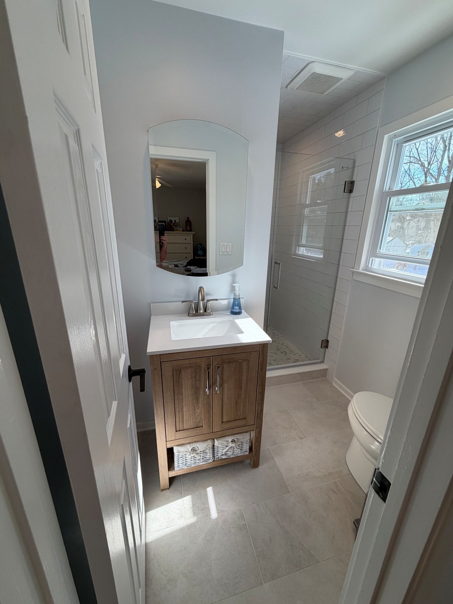 A small bathroom featuring a wooden vanity, a white sink, an oval mirror, a glass-enclosed shower, and a toilet.