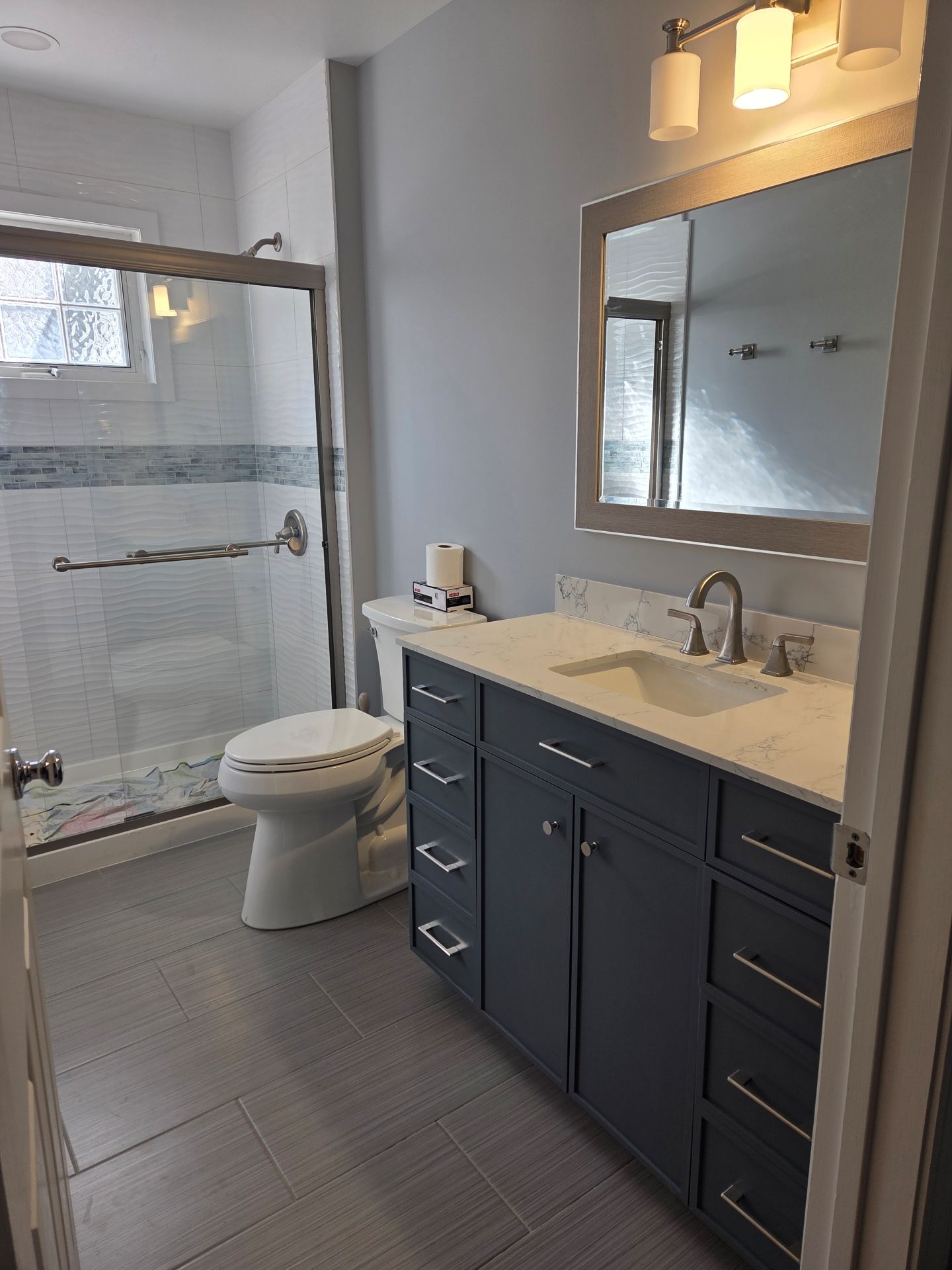 A bathroom with gray vanity cabinets, a white countertop, a mirror, and a walk-in shower with a glass door.