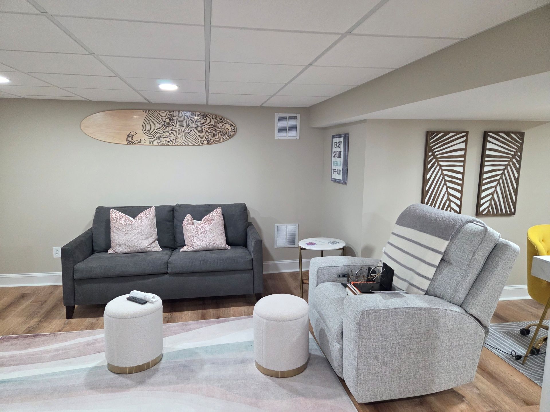 A living room with a charcoal gray sofa, light gray recliner, two round stools, and wall art including a surfboard piece.