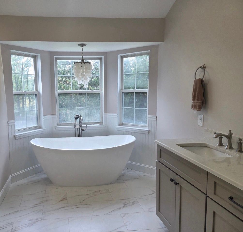 A modern bathroom features a white freestanding tub in a bay window, marble flooring, and a taupe-colored vanity.