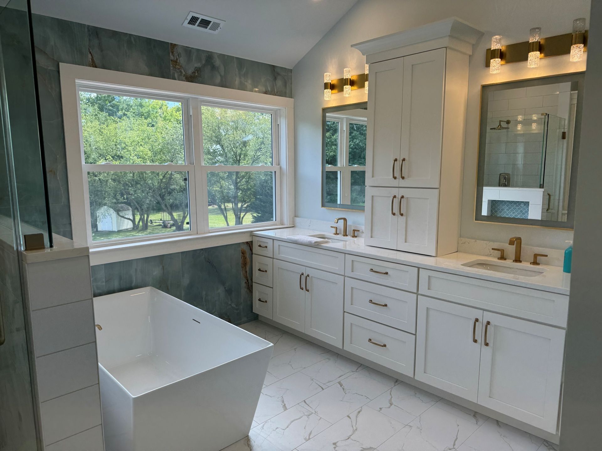 A modern bathroom with a freestanding white tub, double vanity with white cabinets, marble floors, and large window.