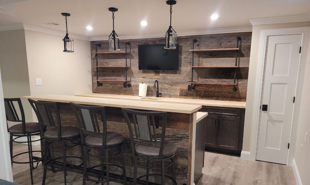 A basement bar with a stone wall, industrial shelving, pendant lights, and four bar stools behind a wooden counter.