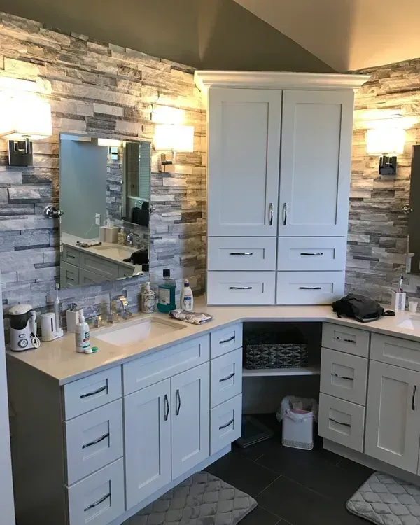 A modern bathroom vanity with white cabinets, a stone-tiled wall, two sinks, and a central cabinet tower in a corner.