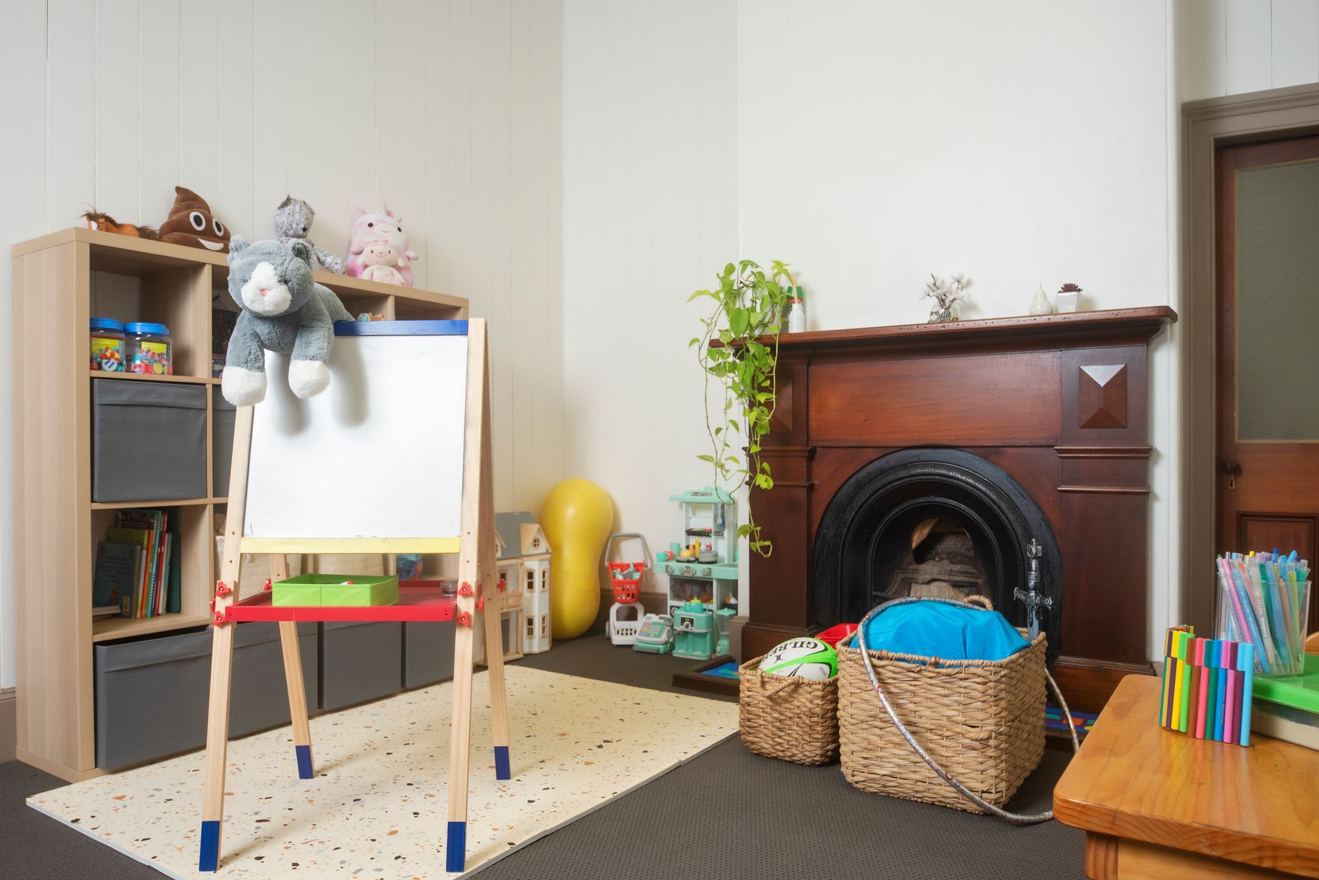 Child's playroom: easel, shelves with toys, fireplace, baskets. White walls, dark carpet.