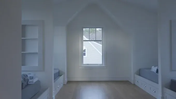 A symmetrical attic room with white walls, a central window, and twin built-in beds with blue bedding on either side.