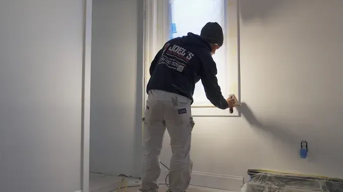 A professional painter in work clothes uses a brush to cut in white paint around a window frame in a room.