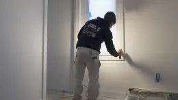 A professional painter in work clothes uses a brush to cut in white paint around a window frame in a room.