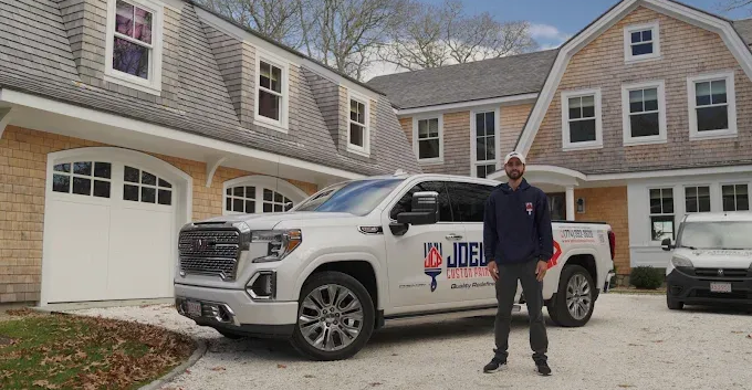 A man stands in front of a white Joe’s Painting pickup truck parked on a gravel driveway before a large shingled home.