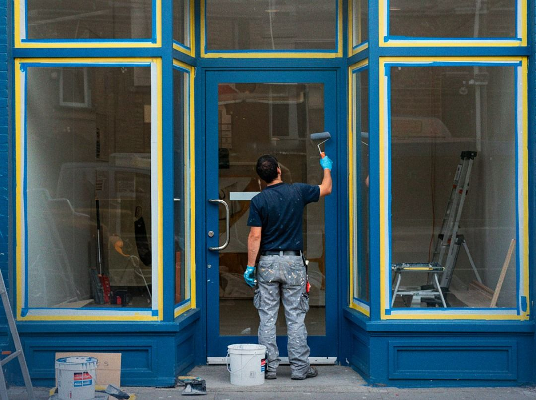 A person in a blue shirt and grey pants painting the blue trim of a storefront glass door and windows.