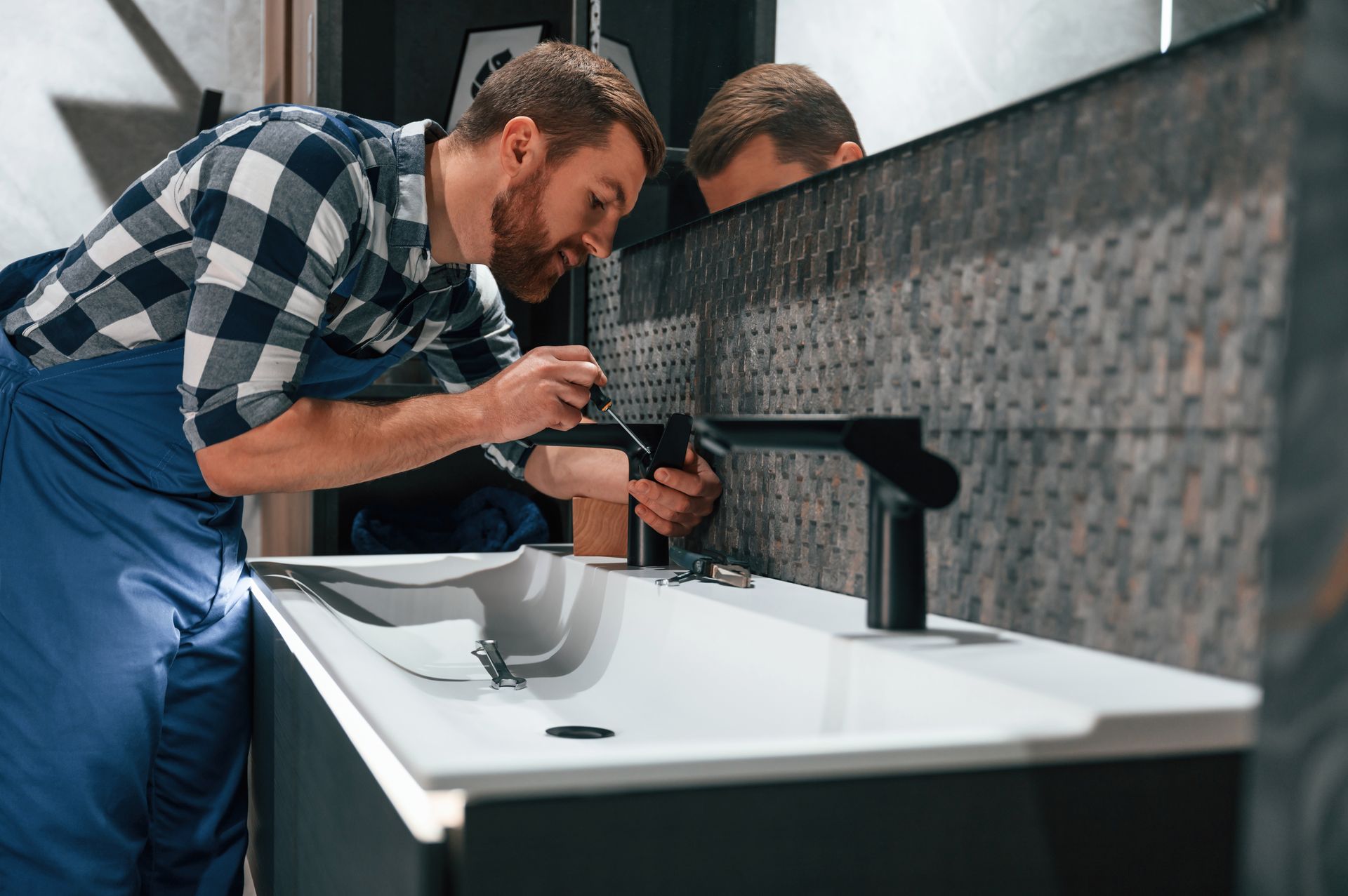 Un plombier professionnel, vêtu d'une chemise à carreaux et d'une salopette bleue, installe un robinet de salle de bain noir et élégant sur un lavabo blanc.