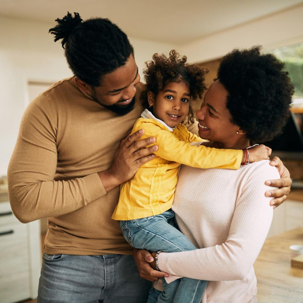 Family sitting on the floor smiling at a toddler. Mom holding child. Dad holding child's hand. Living room setting.